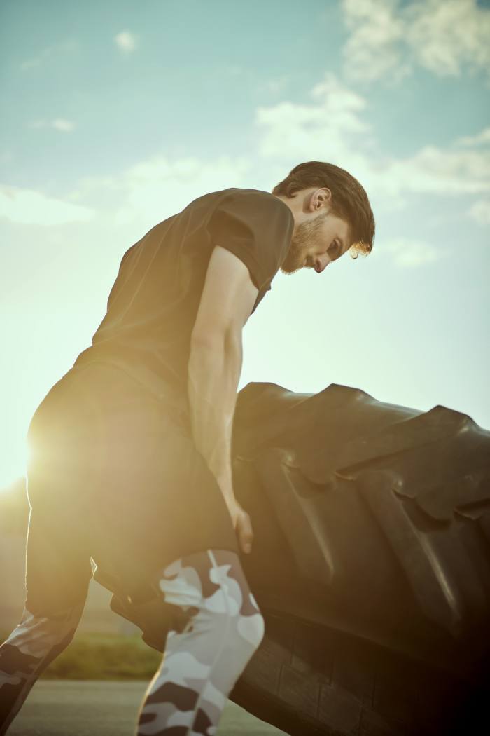 Un tânăr atletic în tricou negru, pantaloni scurți negri și jambiere camuflate ridicând o anvelopă mare de tractor